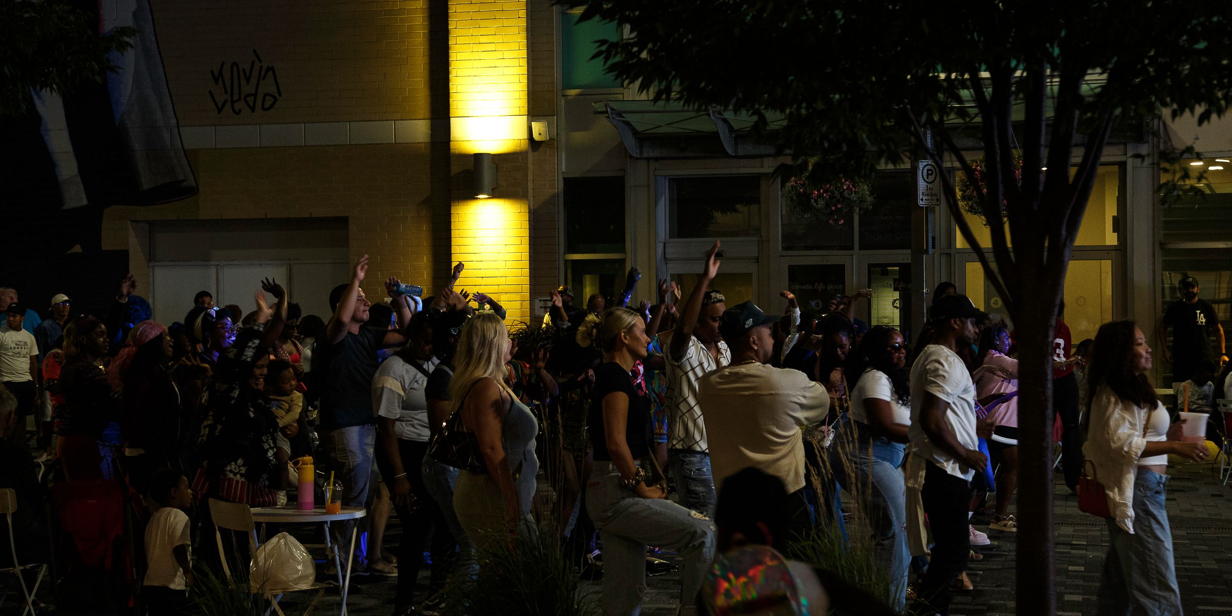 Crowd of people gathered outdoors at night near illuminated building with trees in urban plaza setting.