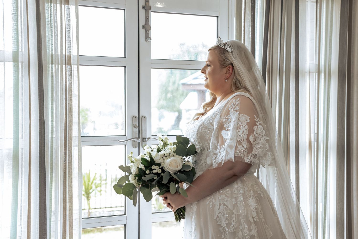 Bride in a lace wedding gown and veil holding a white floral bouquet while gazing out a tall window with sheer curtains.
