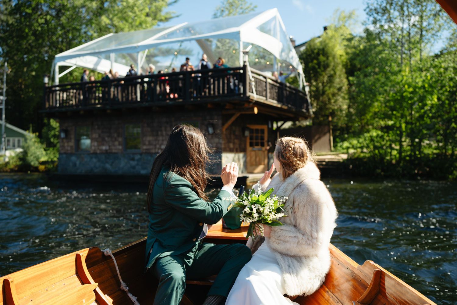 Bride and groom toast champagne on a wooden boat with a glass pavilion venue over a river in the background.