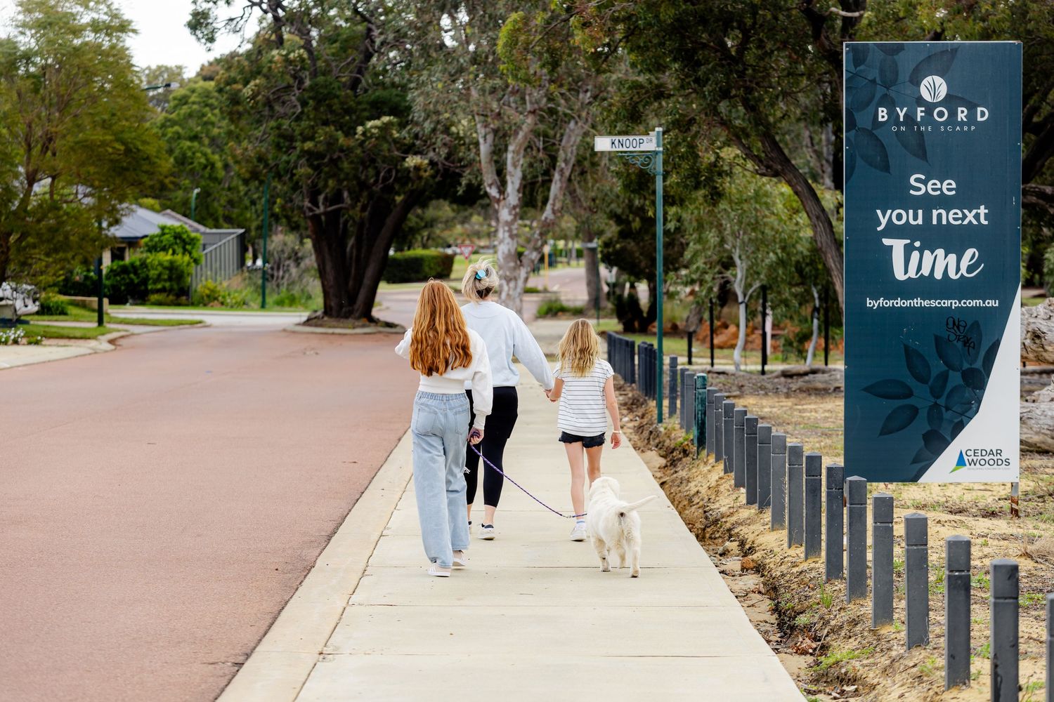 Family walking a white dog on a suburban sidewalk past a Byford town sign reading See You Next Time.