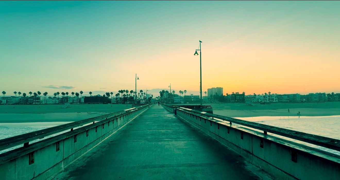 Scenic pier walkway at sunset with warm golden sky, palm trees, and calm ocean waters in the background.