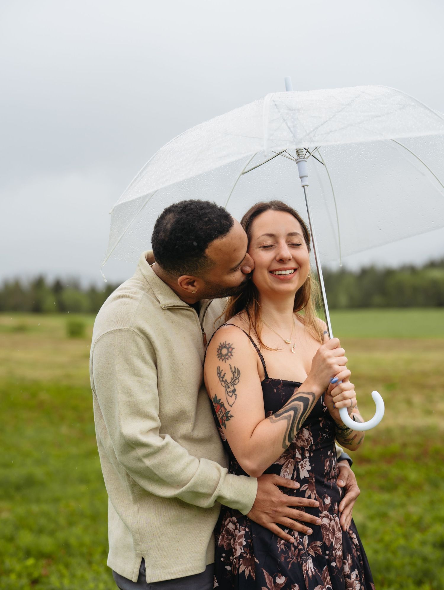 A couple shares a tender moment under a clear umbrella in a green field, the man kissing her cheek as she smiles.