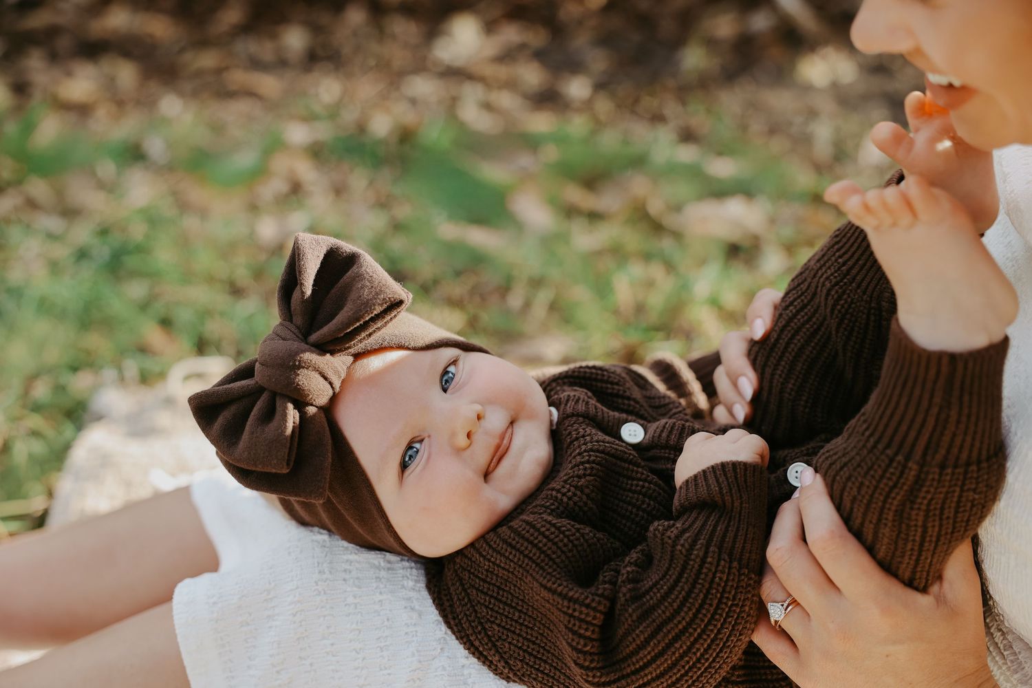 Adorable baby girl in brown knit outfit and bow headband smiling while being held outdoors in fall setting.