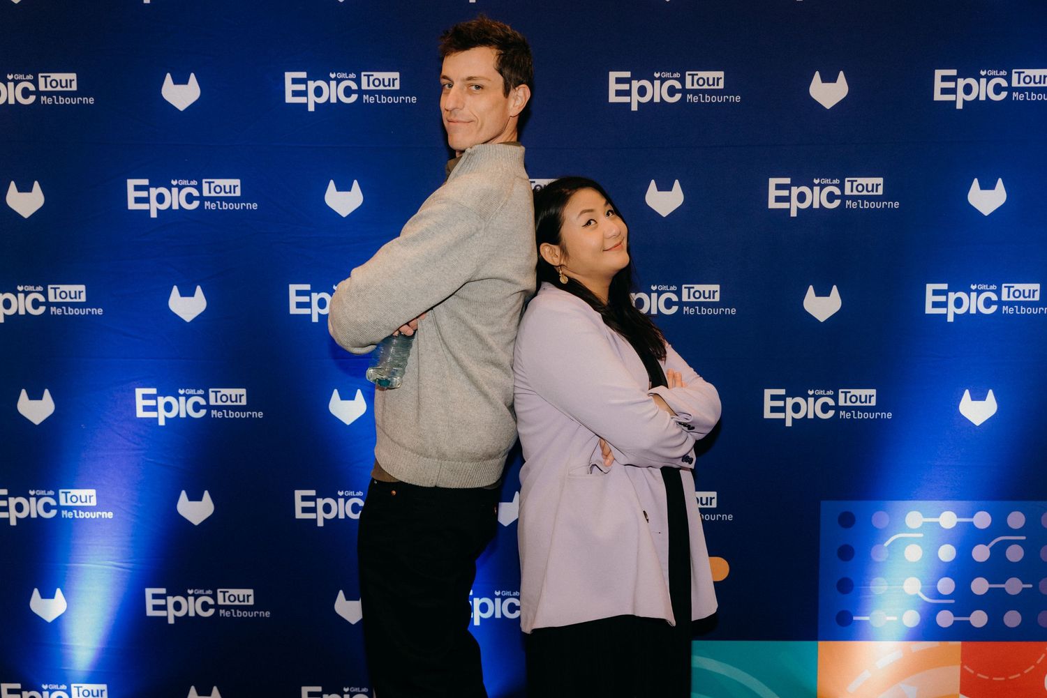 Two people posing back-to-back with arms crossed in front of an Epic blue branded step-and-repeat backdrop.