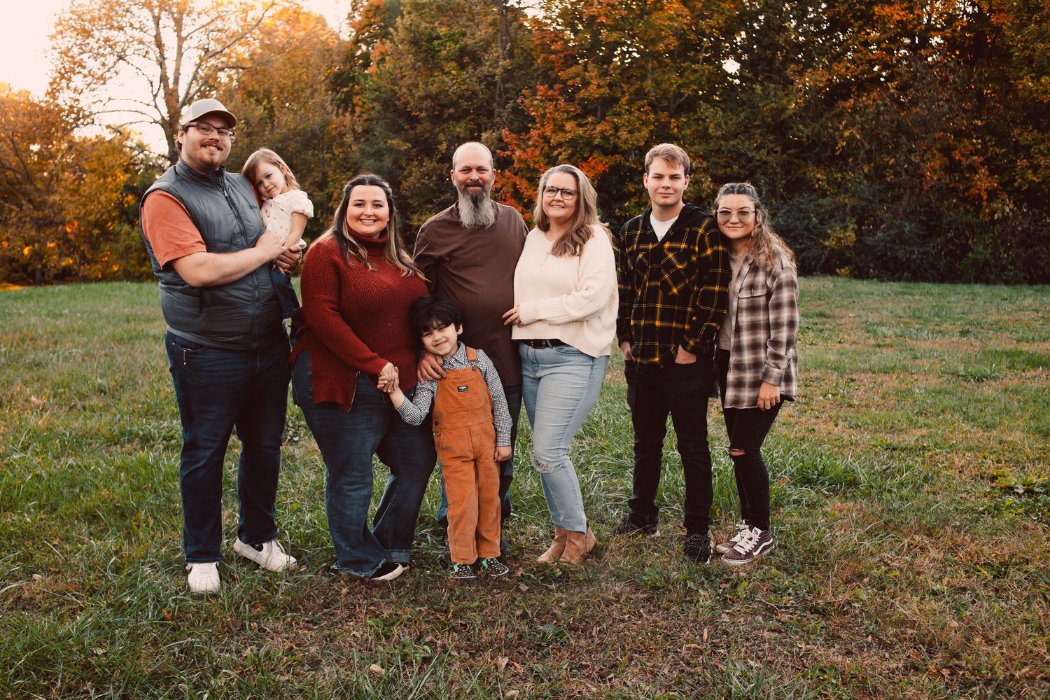 Family of seven posing together in a grassy field surrounded by colorful autumn foliage during golden hour.