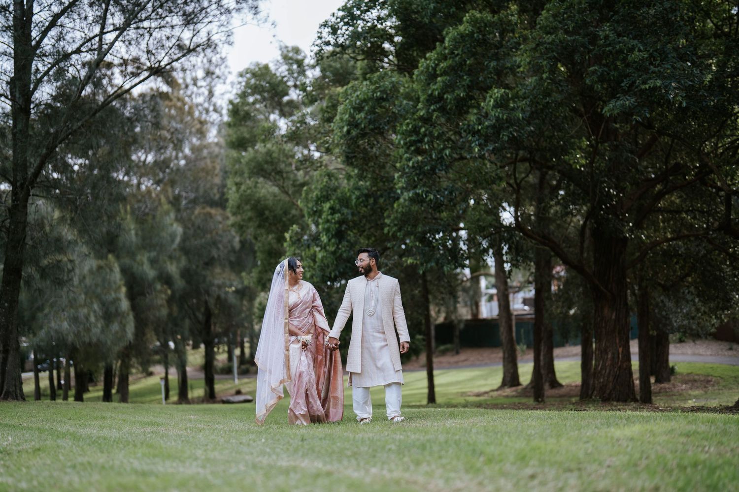 A South Asian couple in traditional wedding attire holds hands while walking across a grassy lawn surrounded by tall trees.