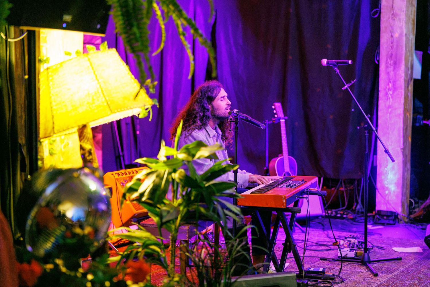 Musician with dreadlocks plays keyboard on a purple-lit stage surrounded by plants, a lamp, and a guitar in the background.
