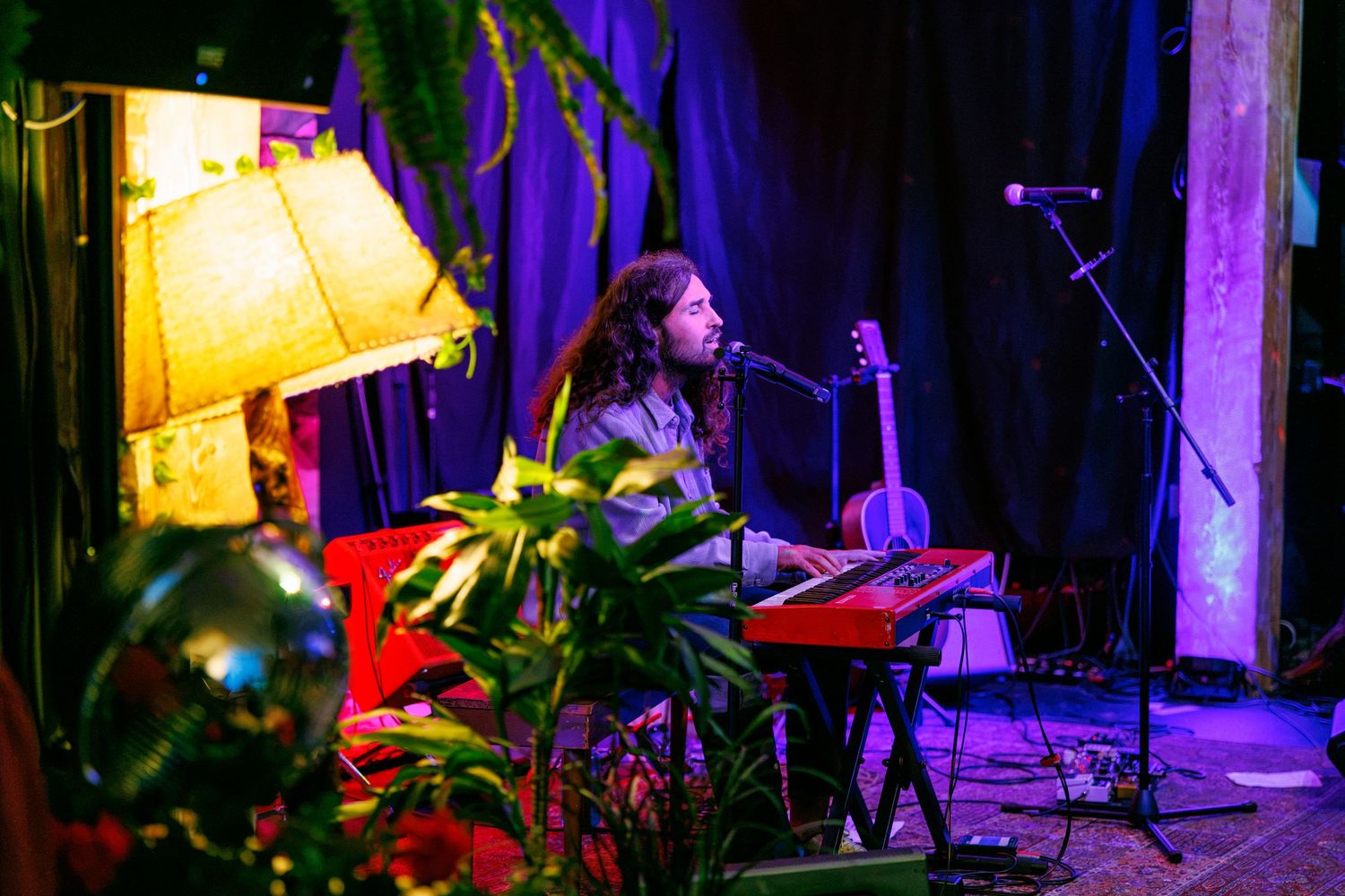 Musician with dreadlocks plays keyboard on a purple-lit stage surrounded by plants, a lamp, and a guitar in the background.