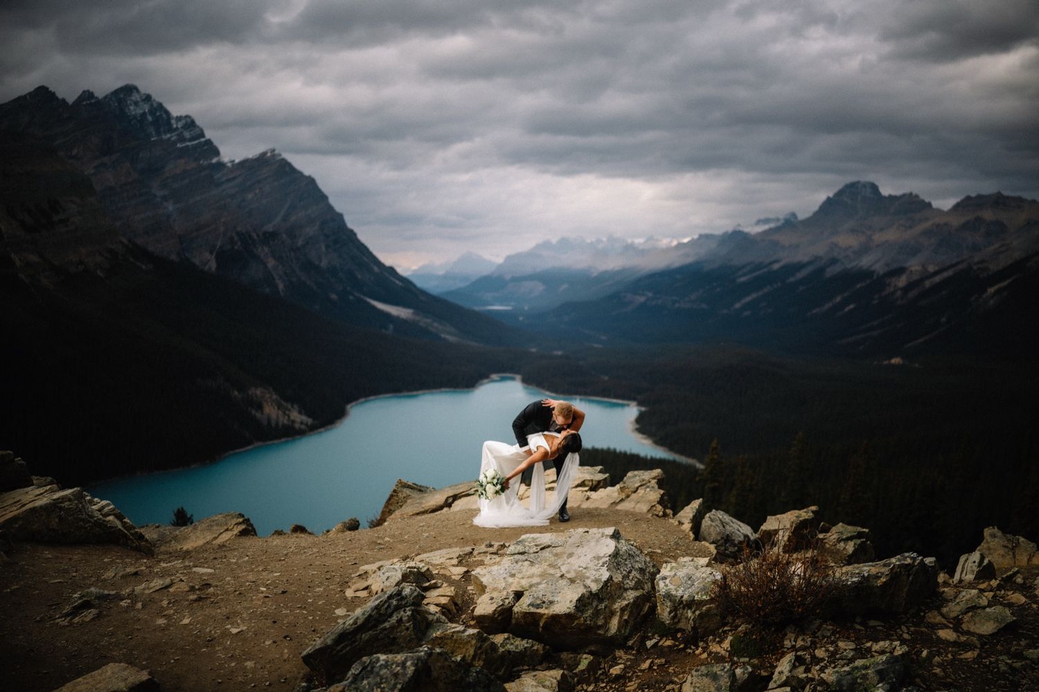 Couple embracing on a rocky overlook above a turquoise mountain lake in the Rockies, wedding photographed by Tkshotz
