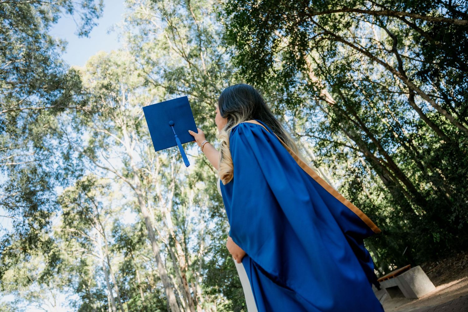 Graduate in blue gown and cap with sunglasses stands before the University of Wollongong Australia sign on a brick wall.