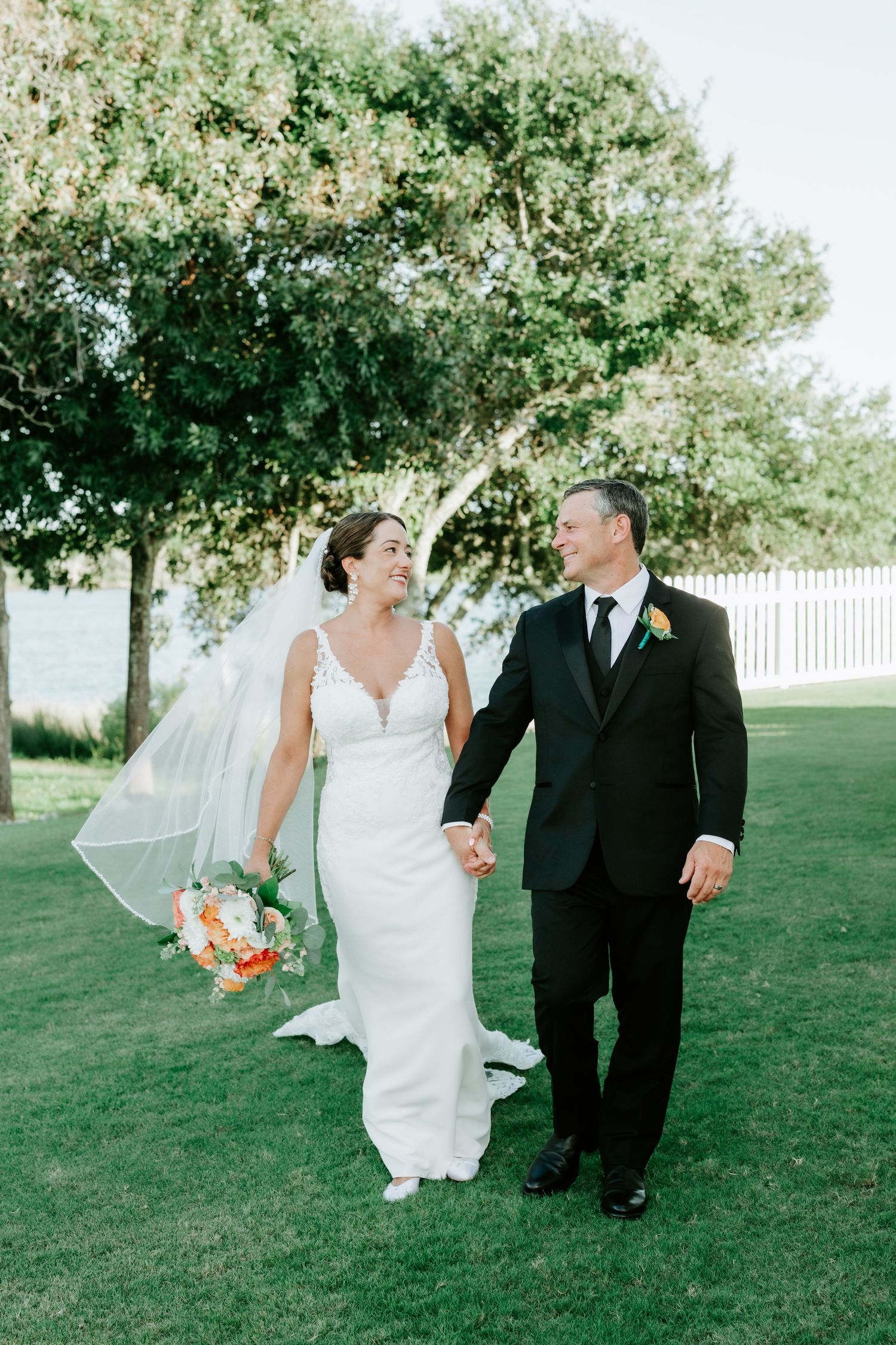 Bride and groom walk hand-in-hand on green lawn, she holds colorful bouquet, veil flowing, trees and water in background.