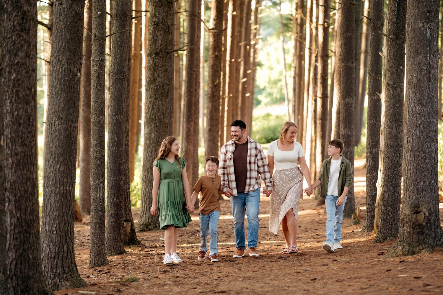 Family of four walking through a pine forest on a sunny day, smiling and holding hands.