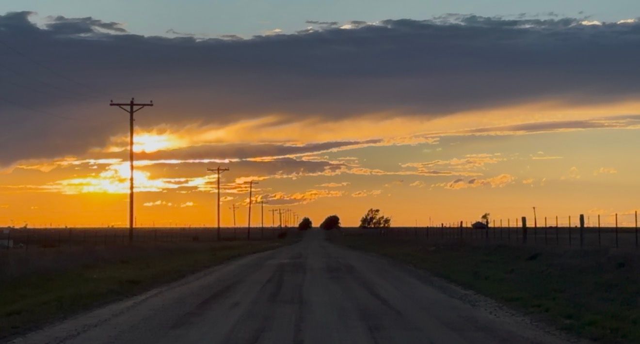 A rural road stretches toward a vibrant golden sunset under dramatic clouds with utility poles lining the flat prairie landscape.