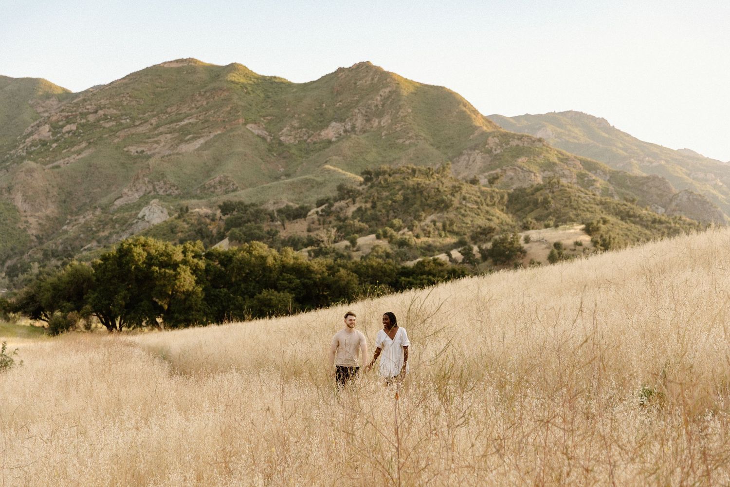 Two people walking through golden dry grass field with mountains and hills in background during sunset.
