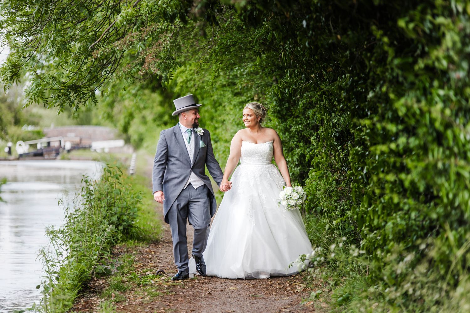 A couple in formal wedding attire walk hand in hand along a peaceful canal path lined with green foliage.