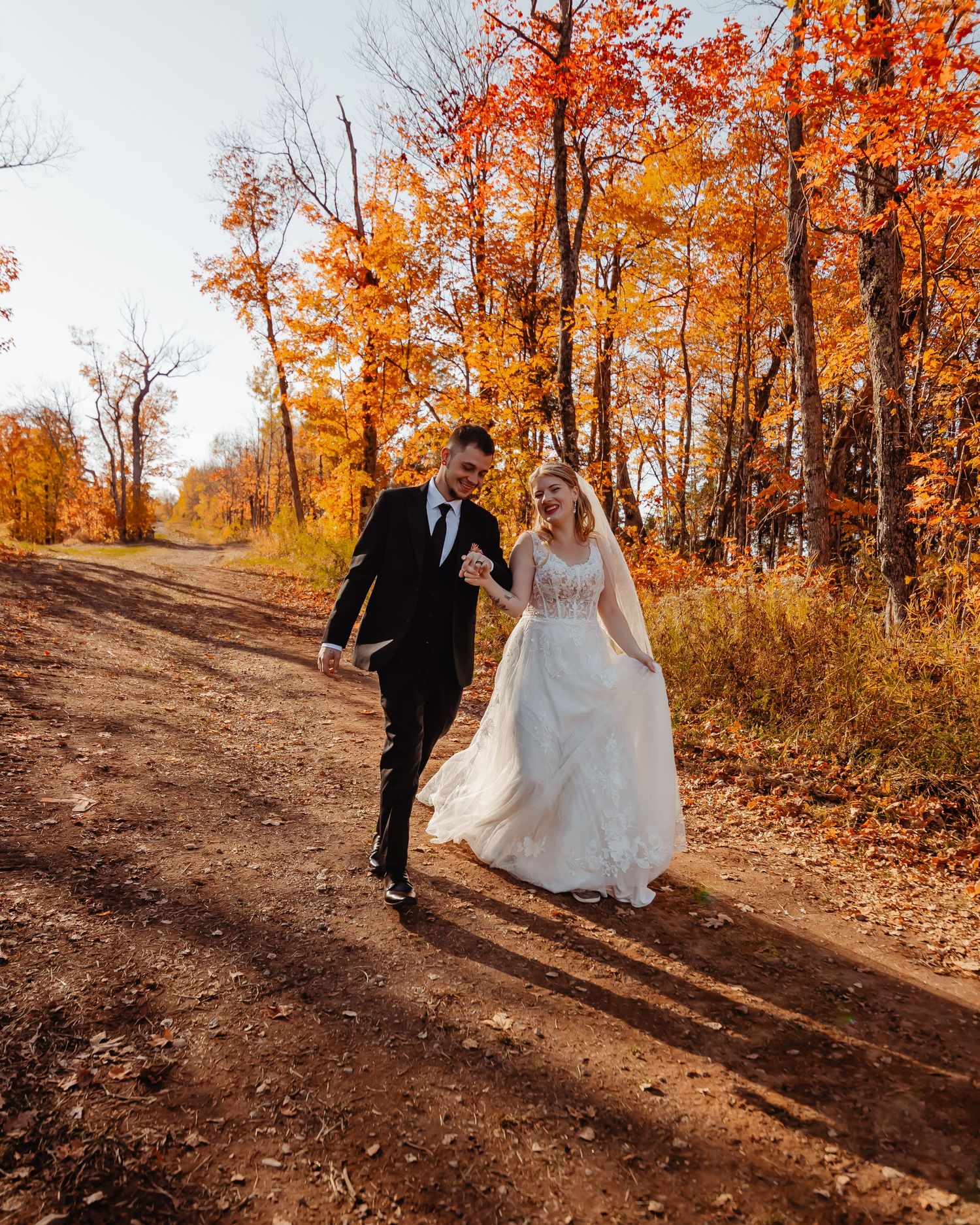 A couple walks hand in hand down a dirt path surrounded by vibrant orange fall foliage.