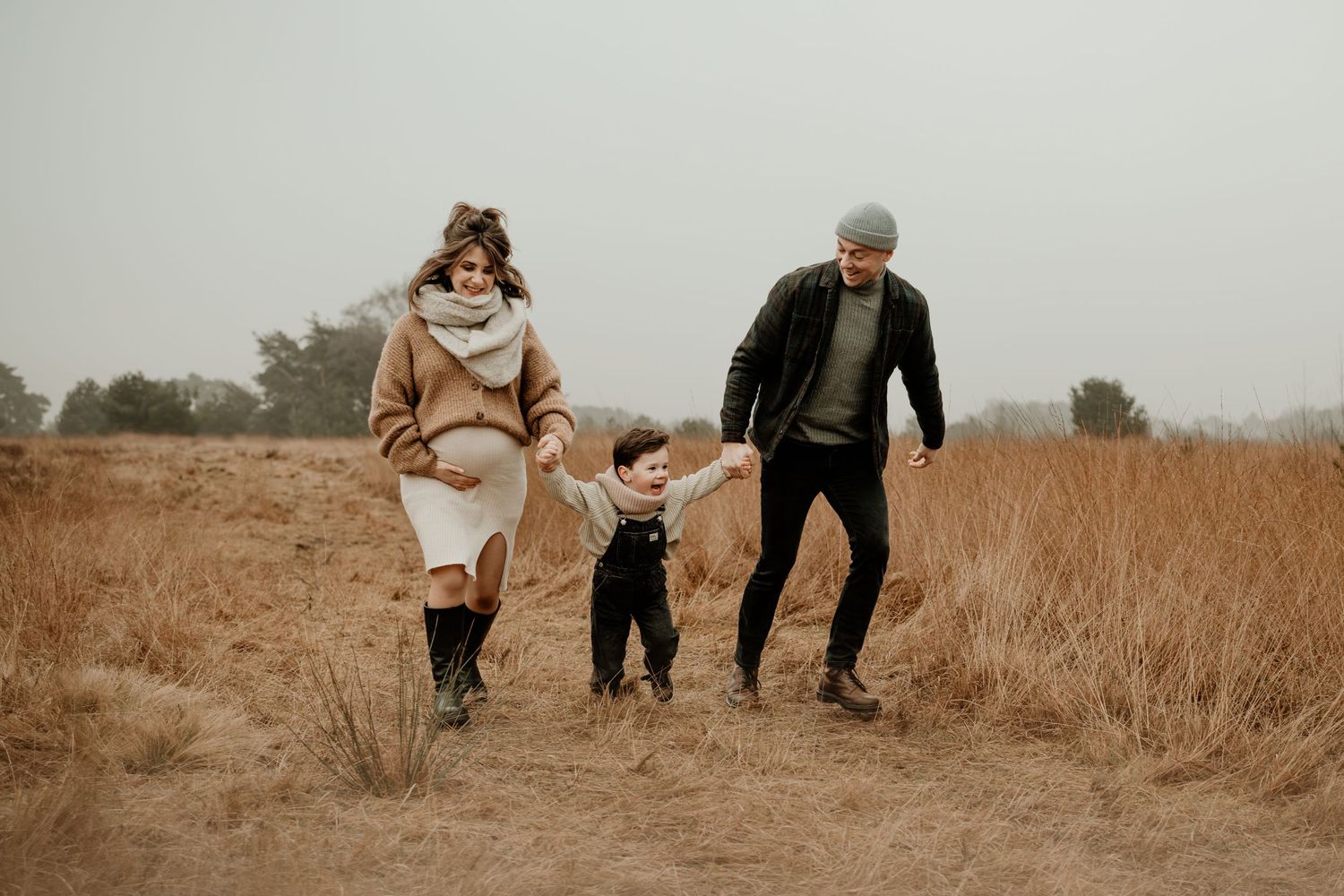 Familieshoot in Geldrop. Een familie wandelt hand in hand door een mistig herfstveld met hoge, verdorde grassprieten op een bewolkte dag.