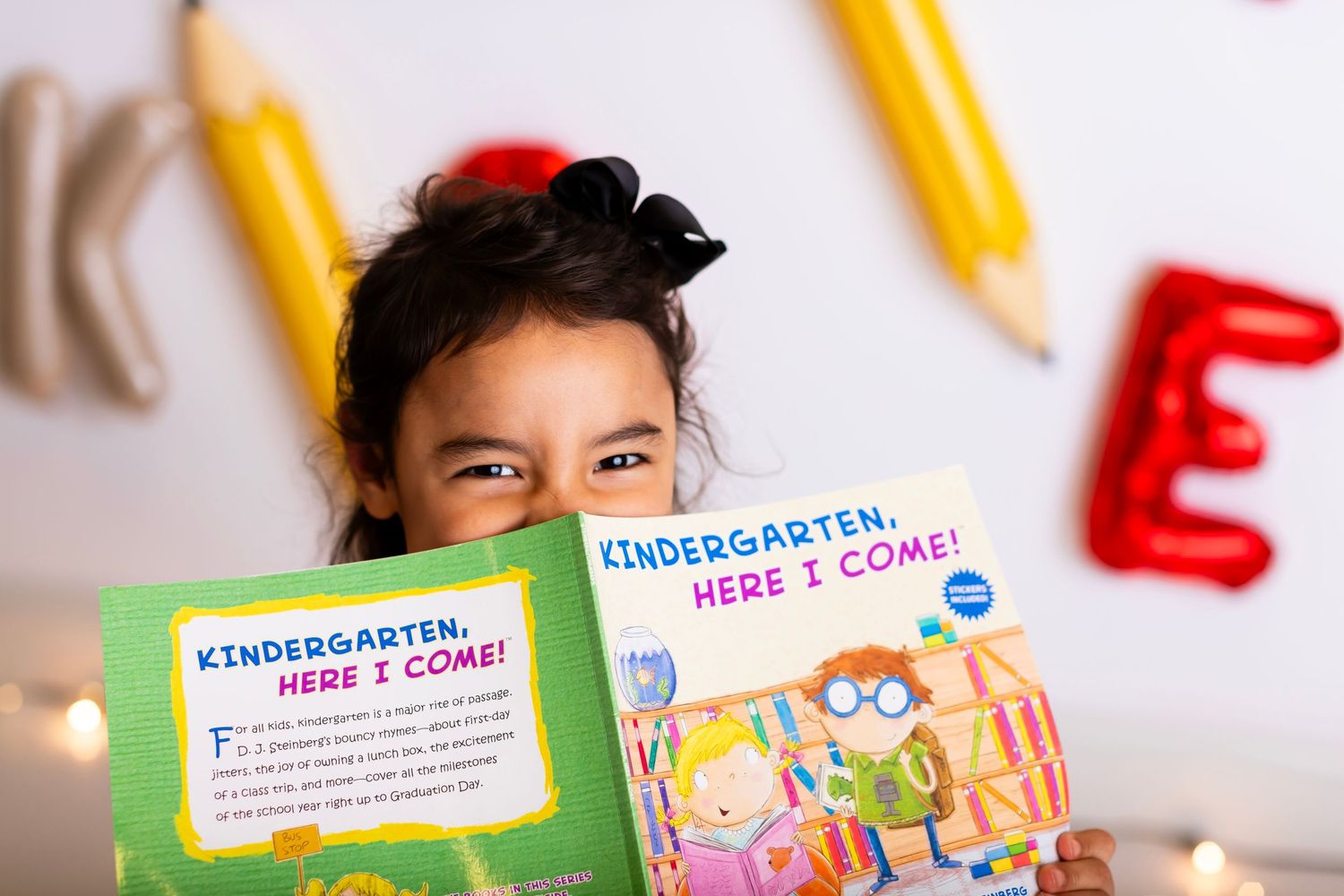 Student smiling while reading kindergarten book with decorative pencils on wall.