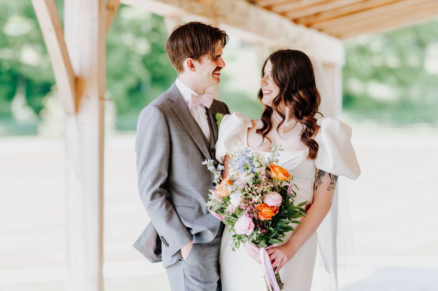 A couple in wedding attire pose together under a covered porch with colorful spring bouquet.
