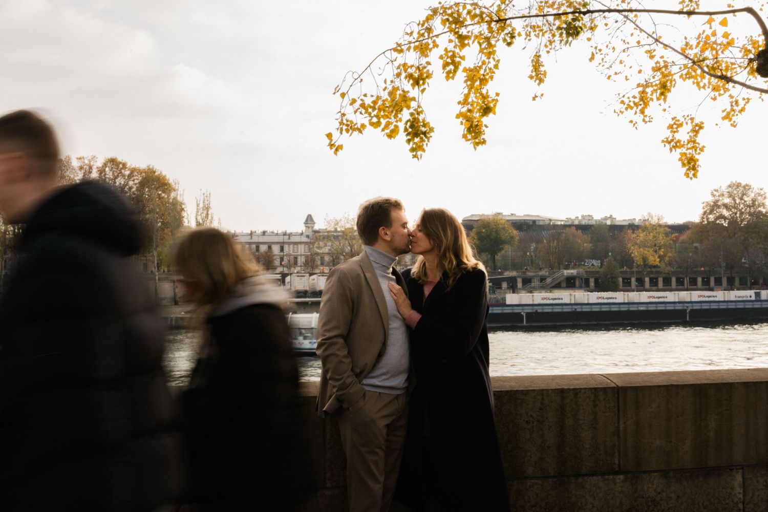 A romantic autumn scene by a riverbank with golden leaves overhead as people stand along the stone wall at sunset.