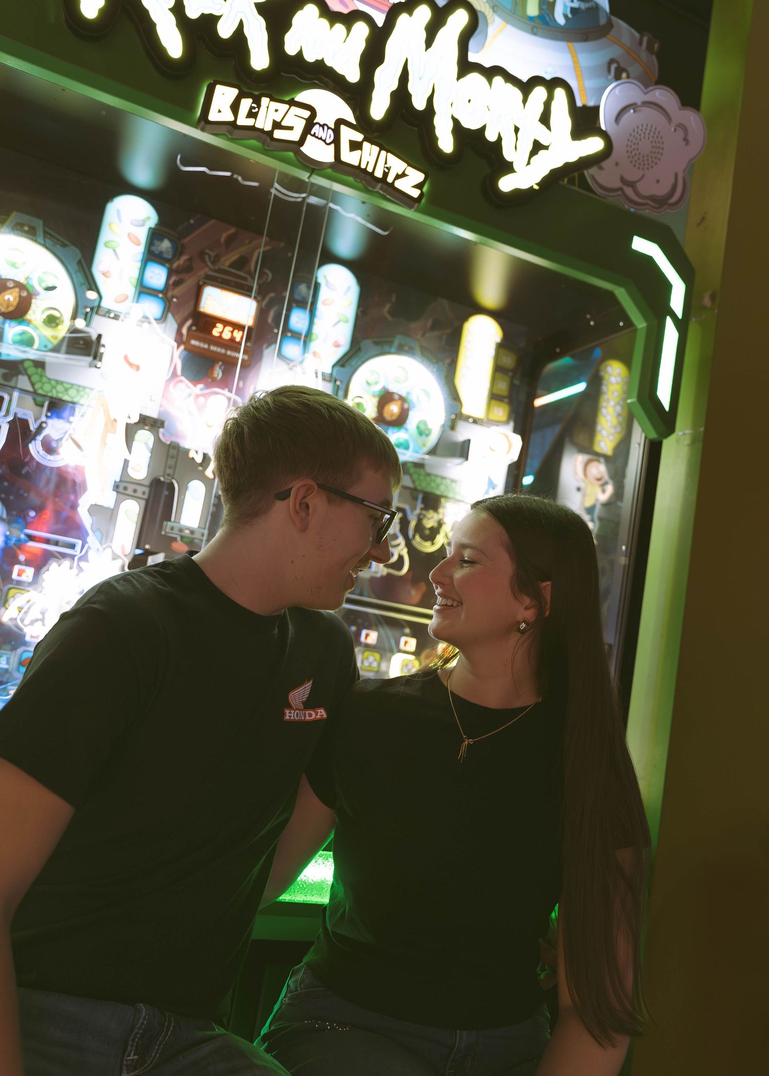 A couple in matching black outfits shares a cozy moment together in front of neon signs at a nighttime venue.