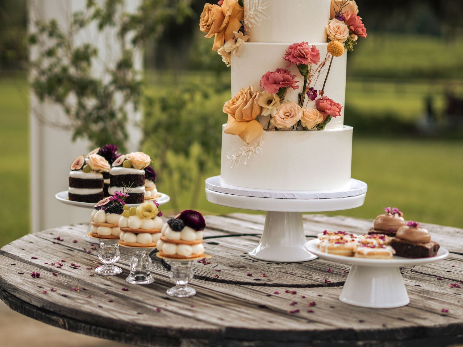 Elegant white wedding cake with peach roses displayed with mini desserts on rustic wooden table.