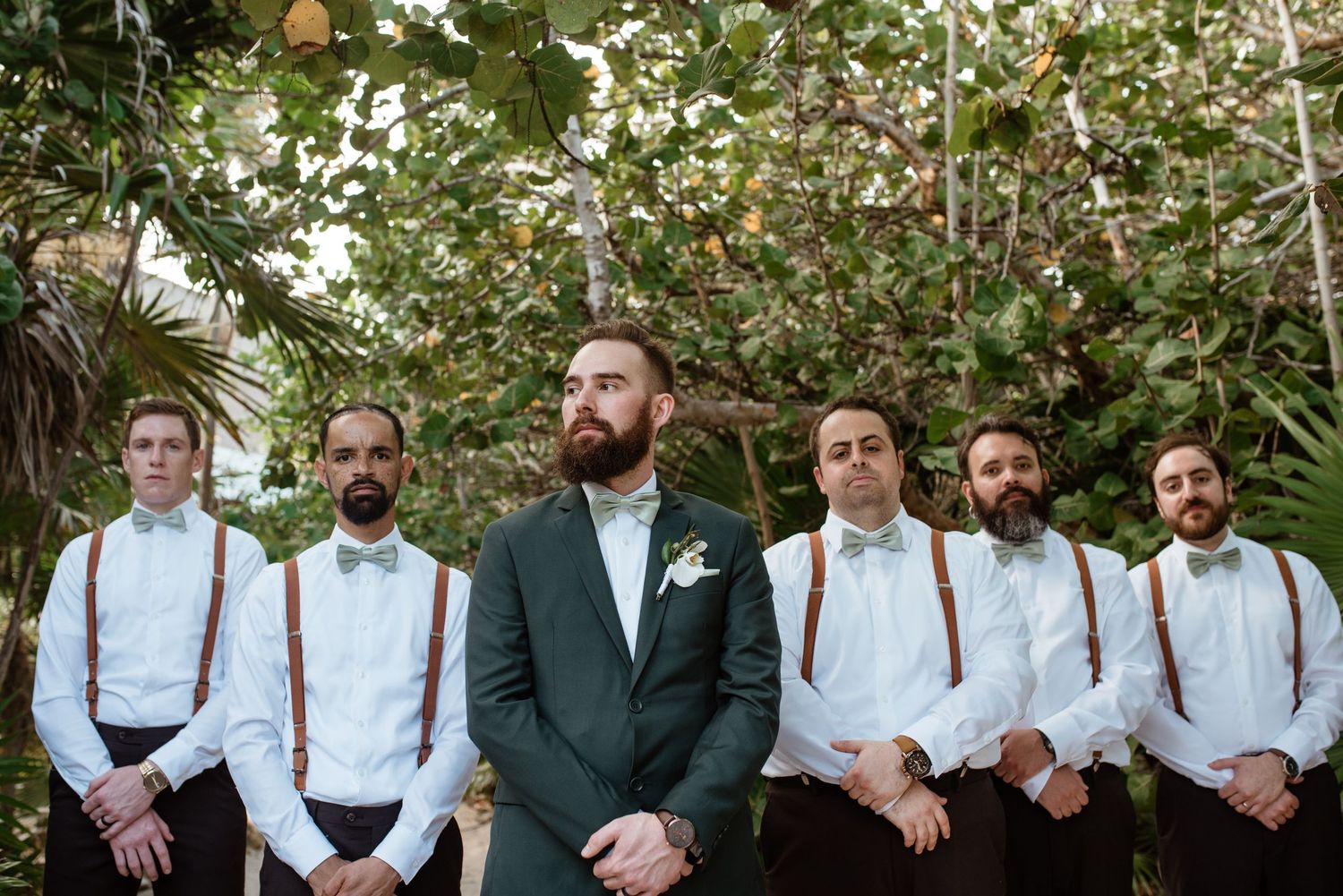 Wedding party lineup with groomsmen in suspenders and white shirts beside groom in dark suit.