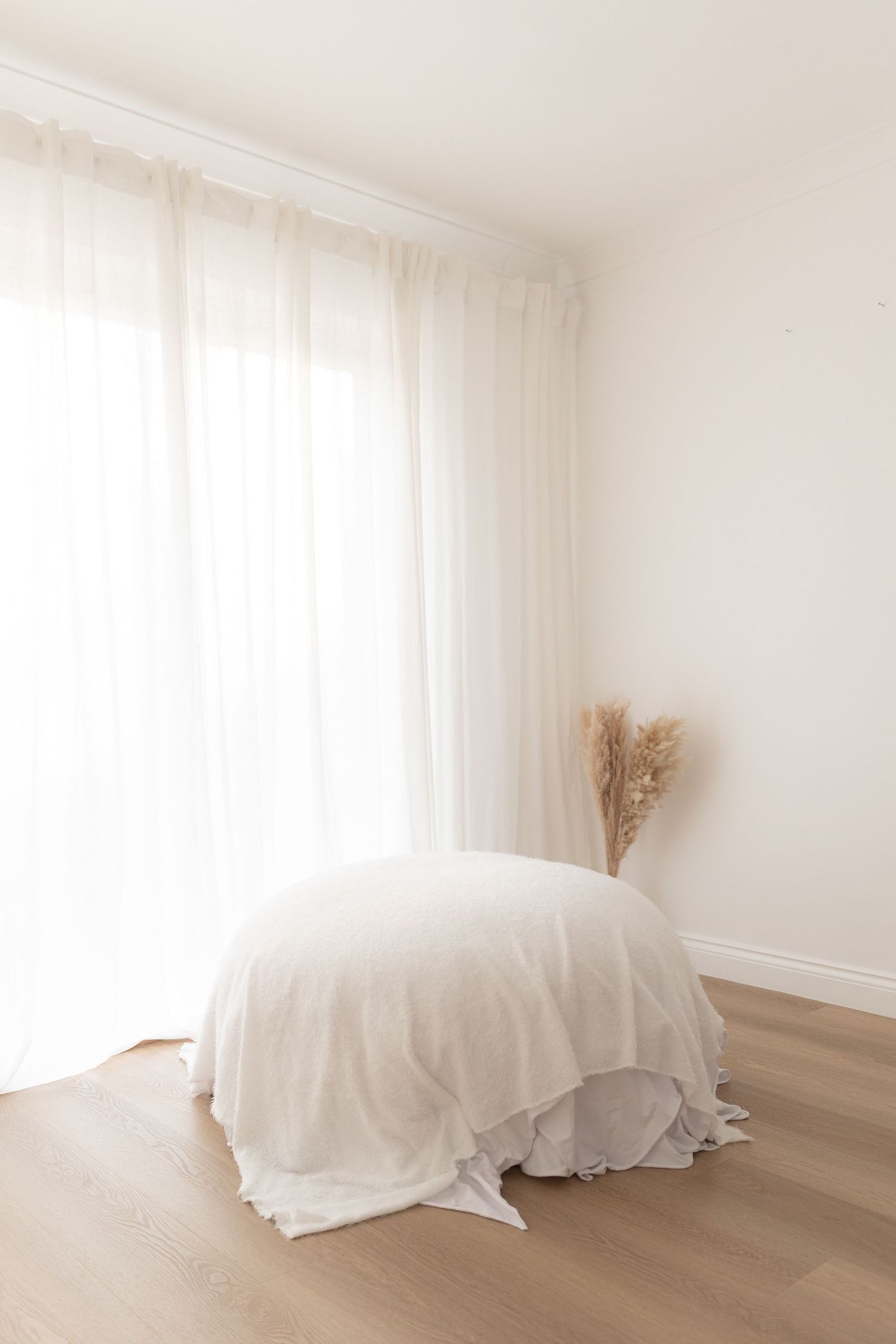 Minimalist room interior with white draping curtains, a draped white furniture piece, and dried pampas grass decoration.