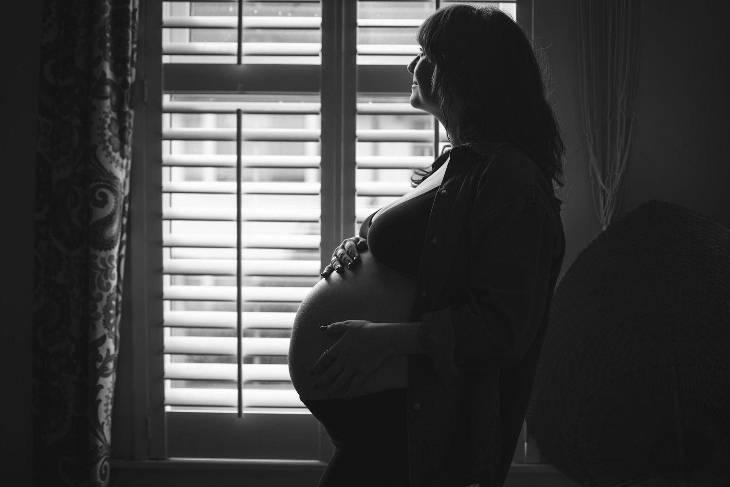 A silhouetted pregnant woman stands by a window with shutters in a moody black and white photograph.