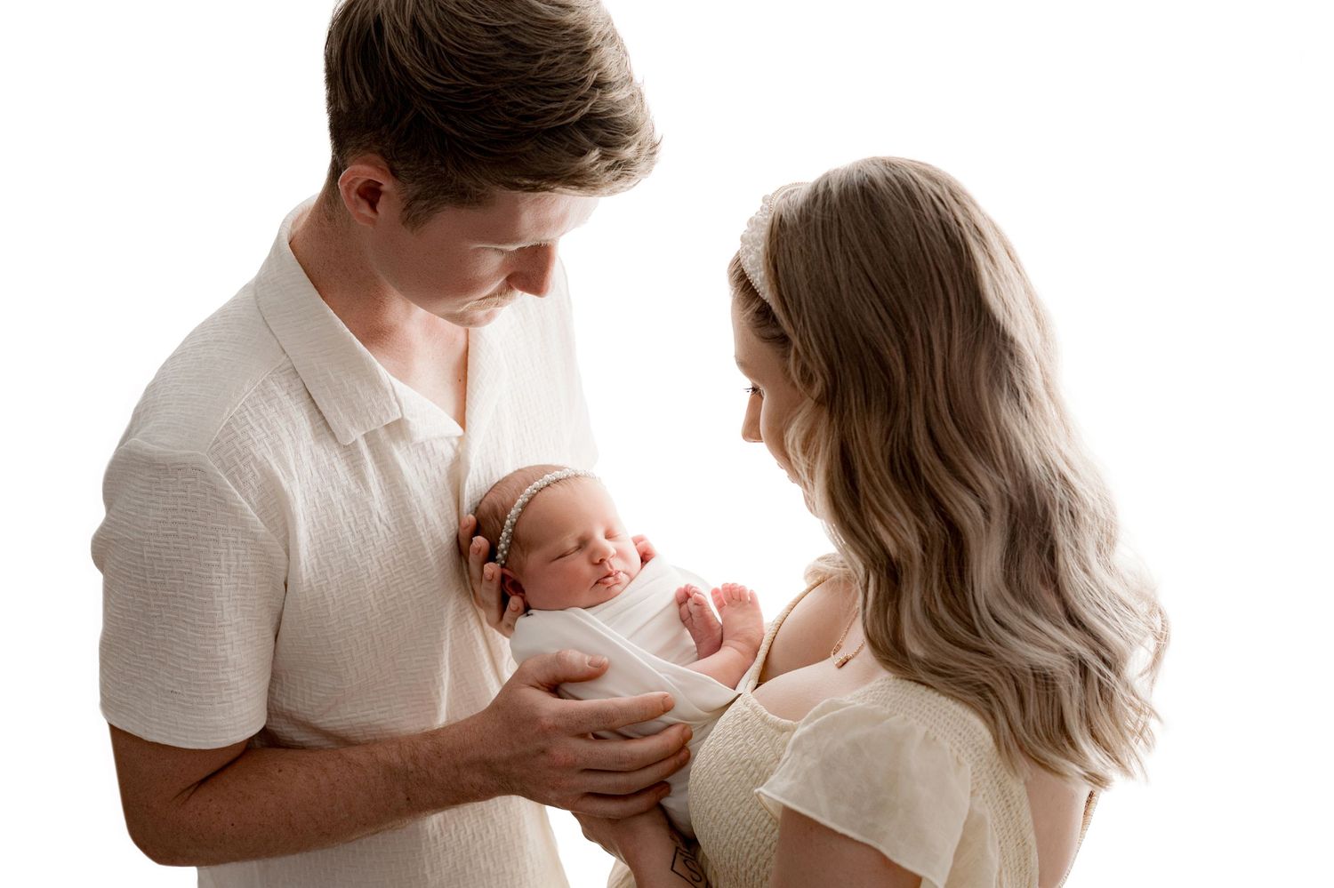 A couple in white clothing holding their newborn baby together in a bright studio setting.