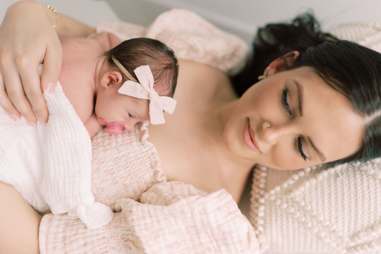 A newborn baby wearing a pink bow rests peacefully on soft bedding while being held in tender embrace.