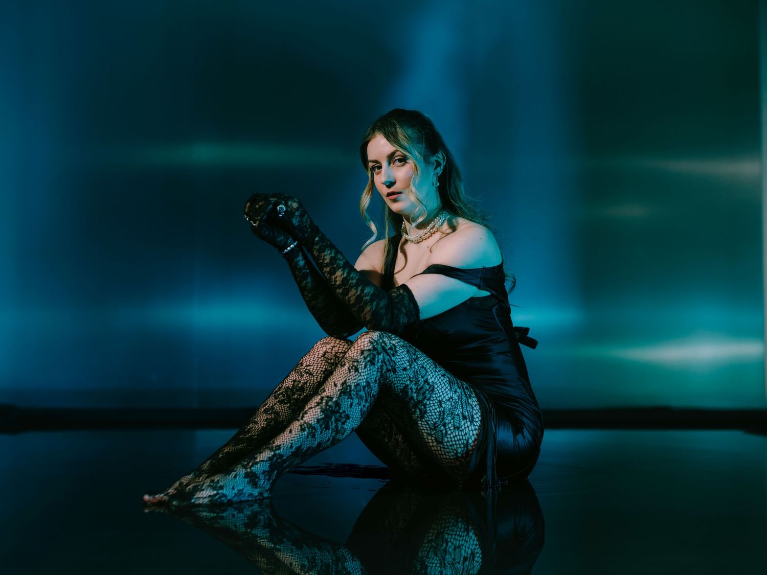 Figure in black lace and blue hair strikes dramatic pose against moody teal backdrop with reflective floor surface.