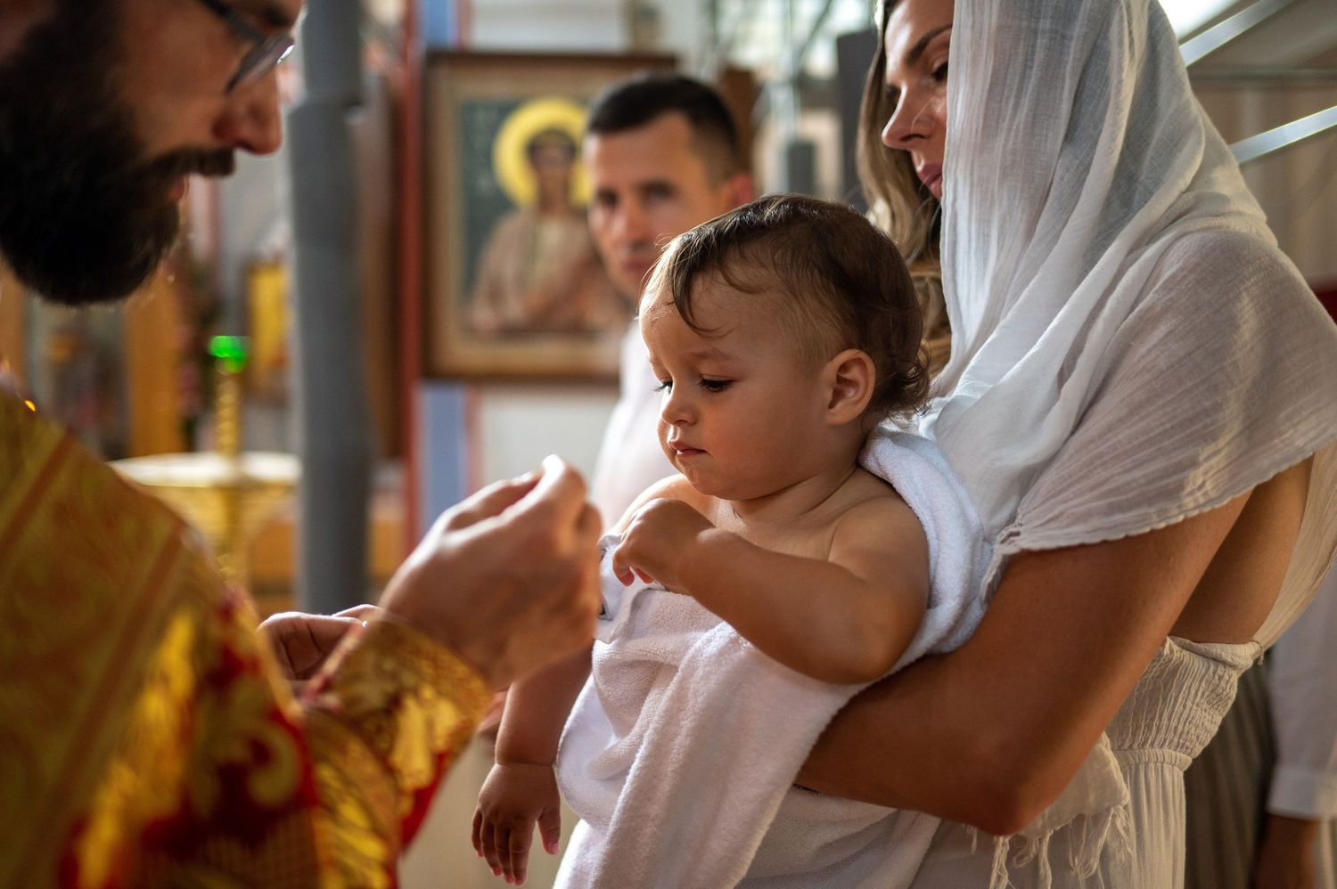 Orthodox Christian baptism ceremony with a baby wearing white being held during the traditional religious ritual.