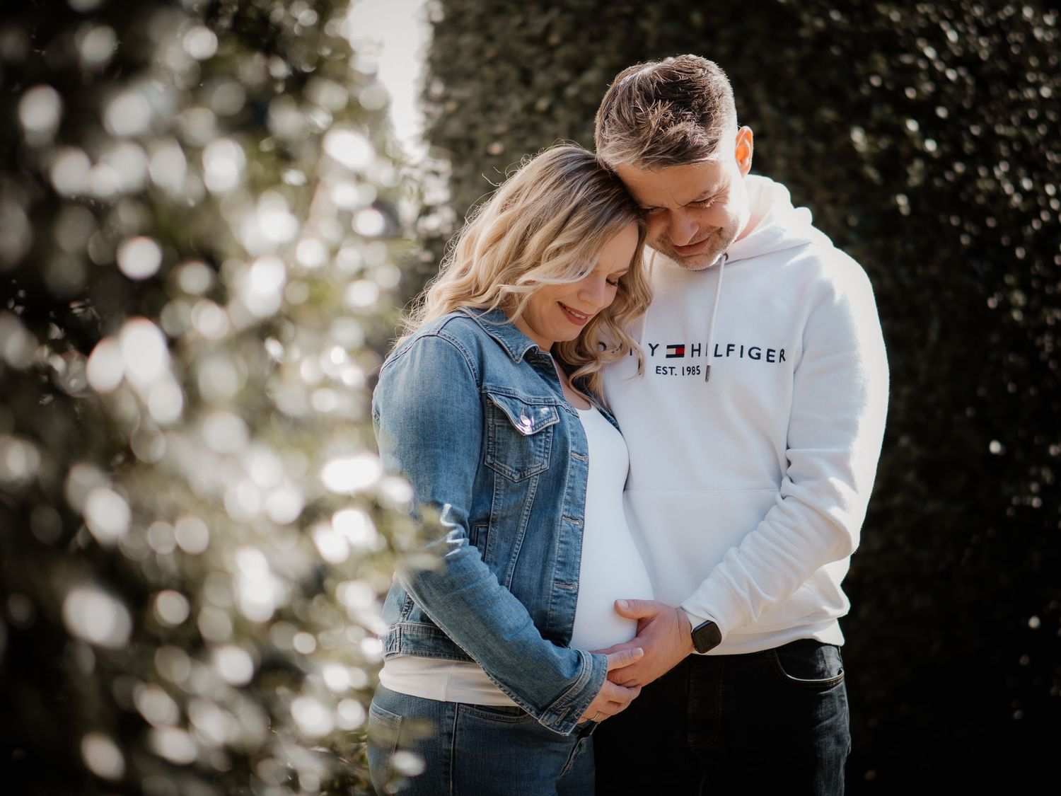 A couple embraces outdoors in a tender moment wearing denim and white hoodie against a bokeh background.