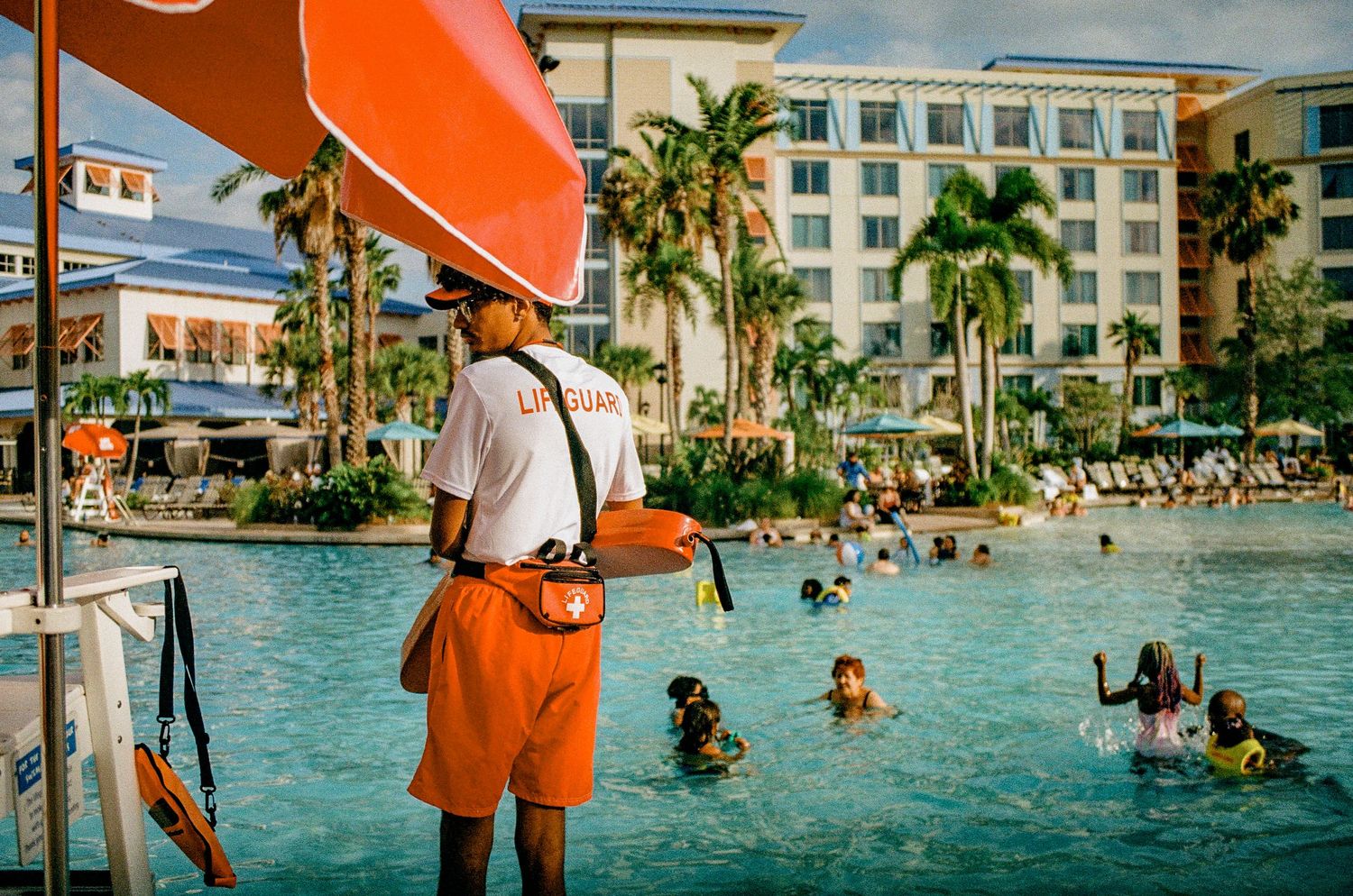 Lifeguard in orange shorts watches over resort pool with palm trees and swimmers.