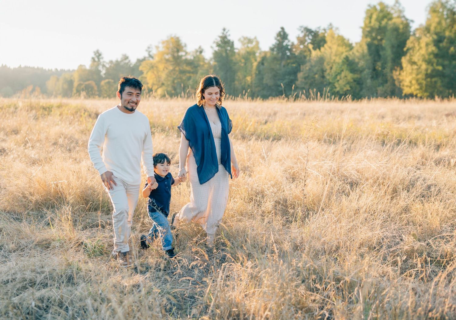 Young family of three walks hand in hand through a sunlit golden grass field outdoors.