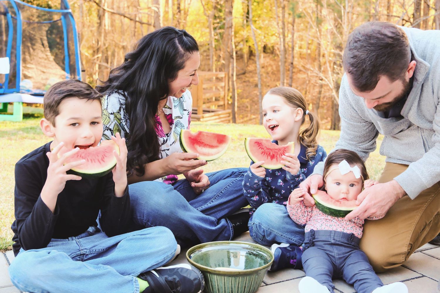 Group enjoys a picnic on blanket outdoors during fall with colorful autumn trees in background and watermelon.