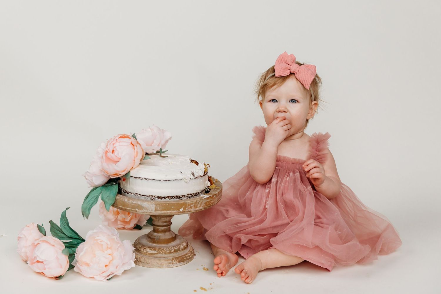Baby in pink dress sitting beside vintage cake stand with roses for first birthday photoshoot.