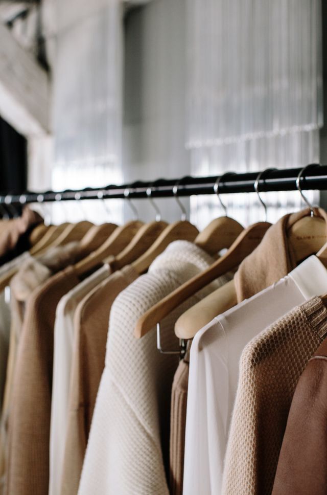 A minimalist clothing rack with neutral beige, brown and white sweaters and tops hanging on wooden hangers.