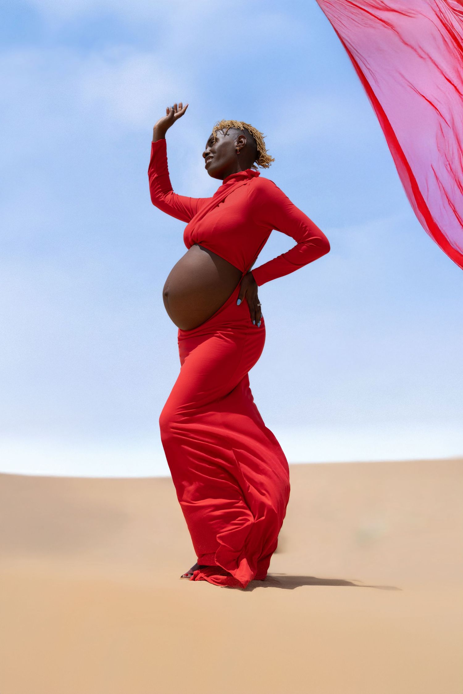 A graceful figure in a flowing red dress poses expressively against sand dunes under a bright blue sky.