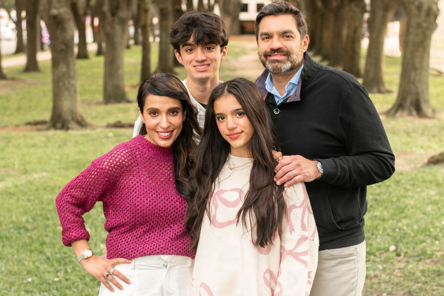 A family poses outdoors in casual attire against a backdrop of trees in a park setting.