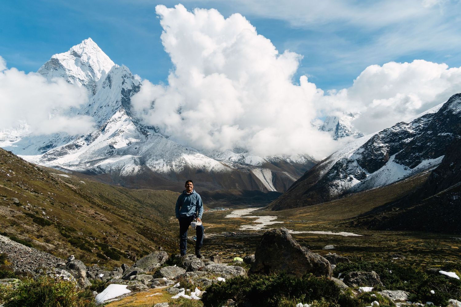Hiker stands on mountain trail with snow-capped peaks and dramatic clouds in the background.
