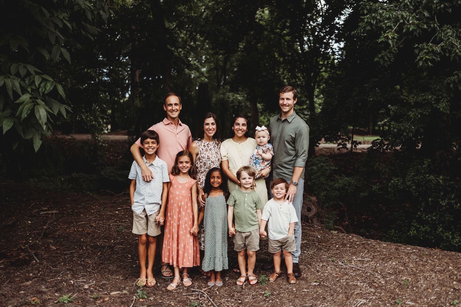 A group photo of a mixed family with children standing together in a wooded area.