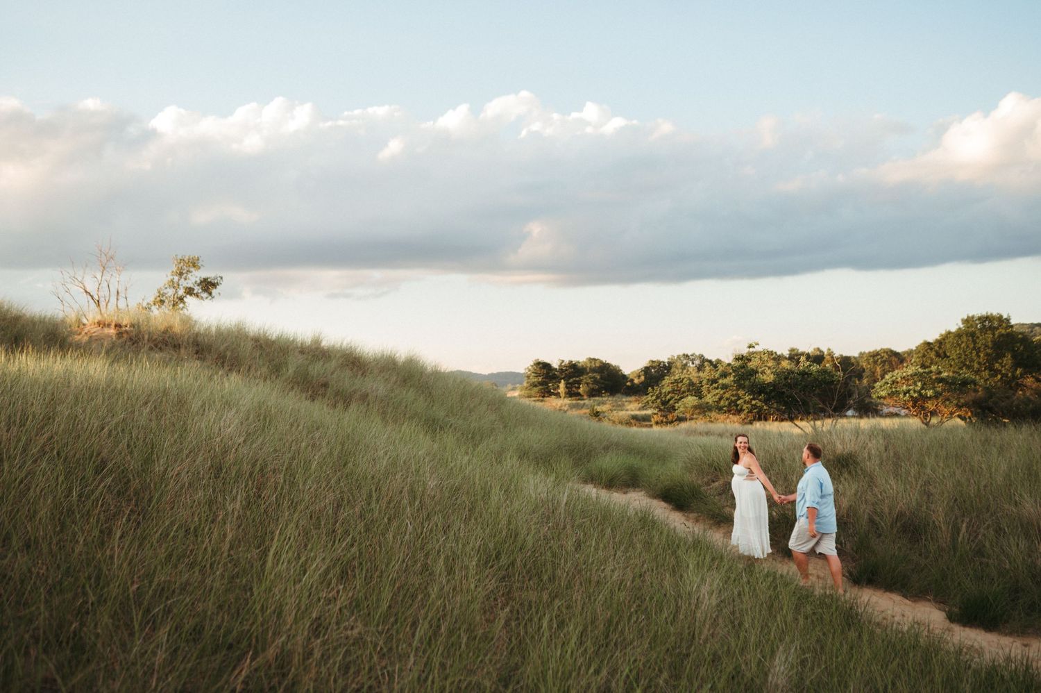 Two people in light colored clothing walk along a grassy coastal path at sunset with clouds overhead.