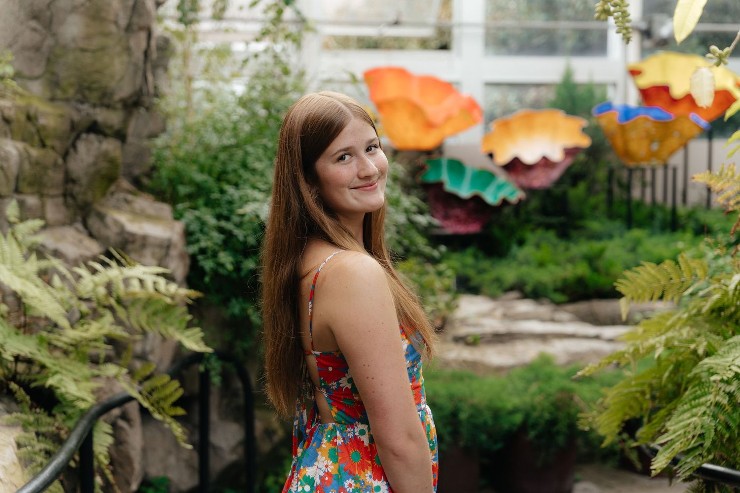 Someone in a floral dress stands in a garden setting with colorful glass flower sculptures in the background.