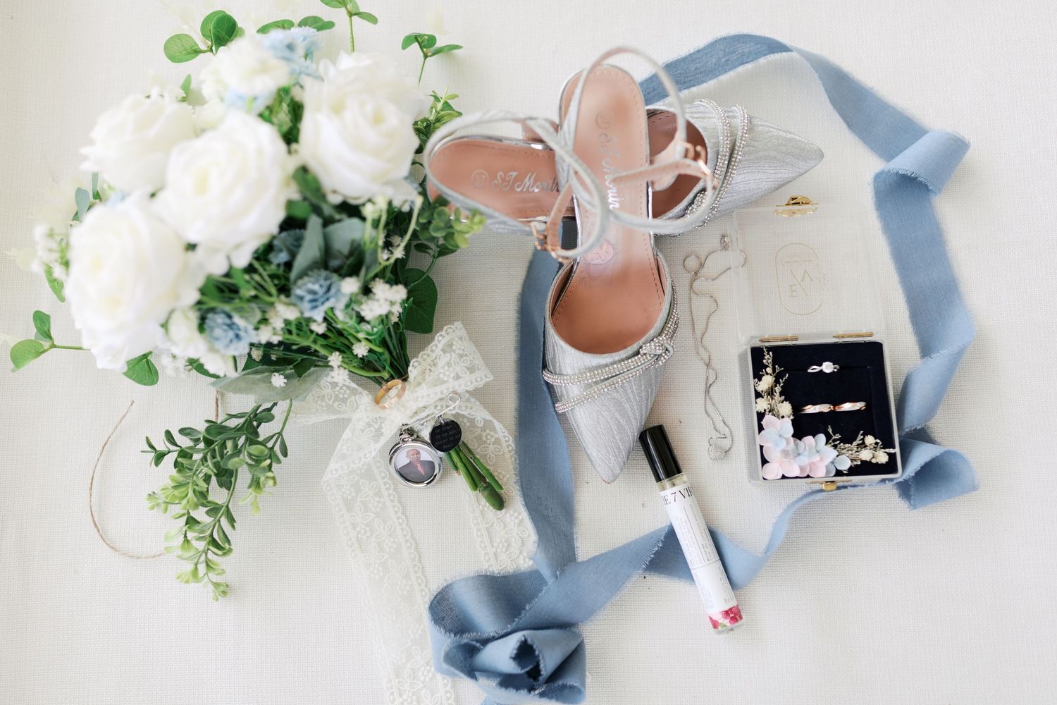 Wedding details flatlay with white flowers, blue ribbon, and silver shoes.