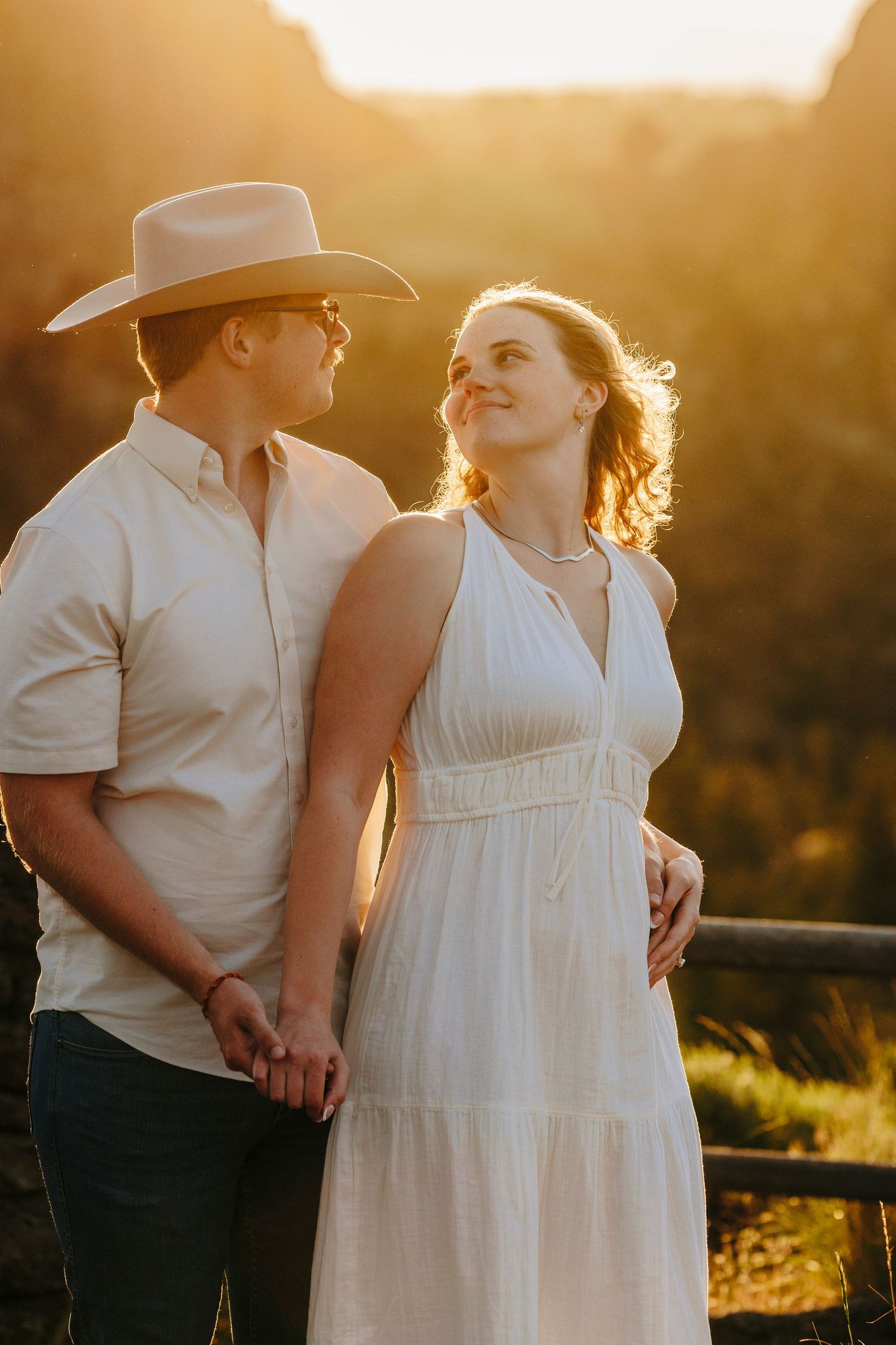 Romantic couple in western attire embracing against golden sunset light by wooden fence at golden hour.