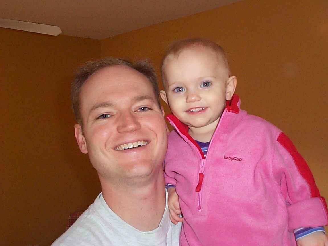 A happy parent and small child in a bright pink jacket smile together for a close-up photo indoors.