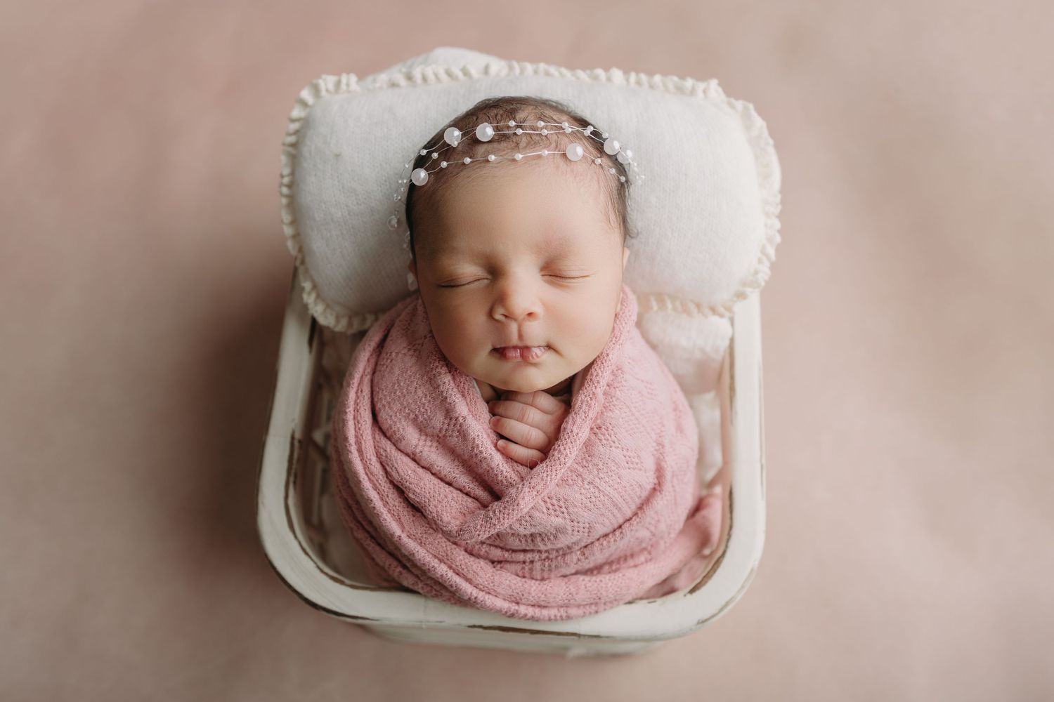 Sleeping newborn swaddled in pink wrap lies peacefully in white cushioned box wearing delicate headband.