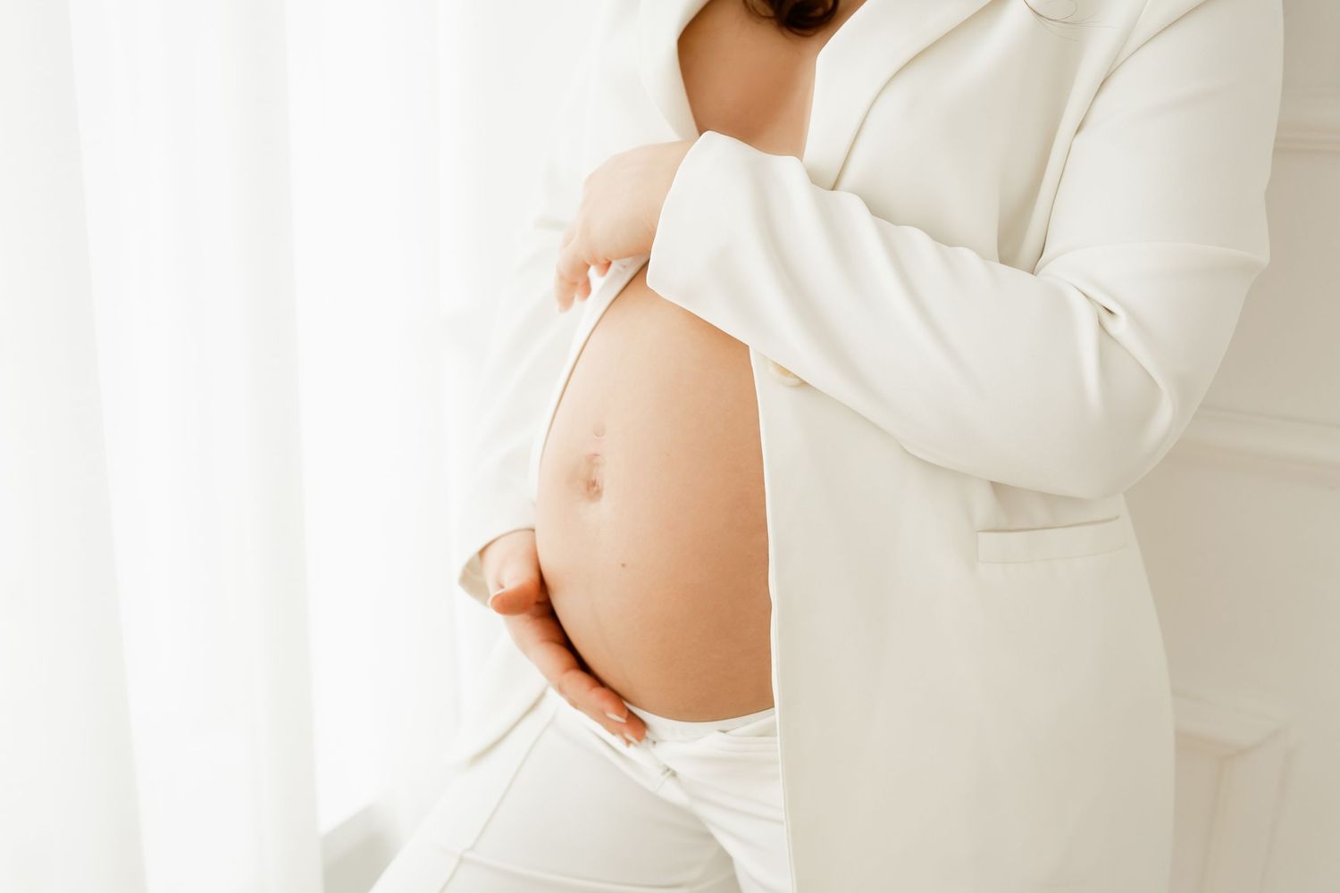 Side view of a pregnant belly in a white sweater against soft window light.